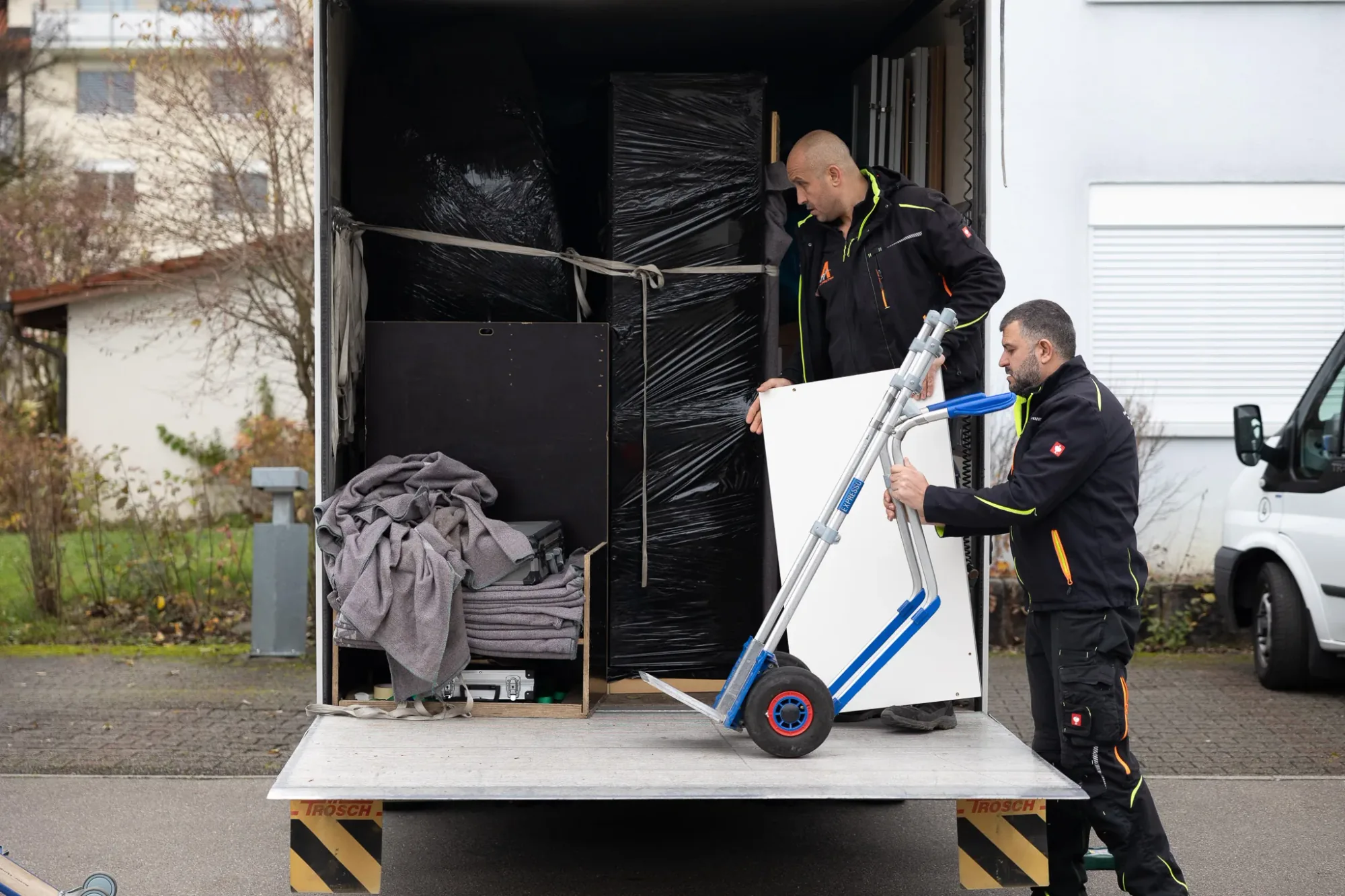 Zwei Mitarbeitende von Activ Umzüge entladen ein verpacktes Möbelstück über die Hebebühne des Umzugsfahrzeugs
